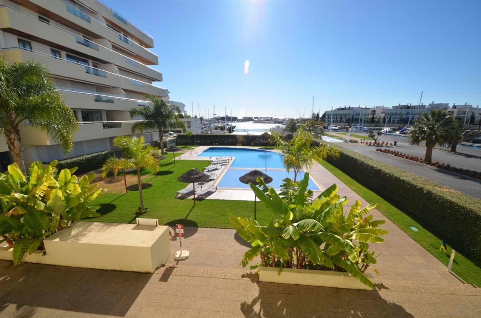 A hotel with a pool area, including lounge chairs and umbrellas, surrounded by greenery and a clear sky.