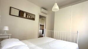 Une chambre à coucher comprenant un lit, une table de chevet avec une lampe, un luminaire suspendu, un miroir et un appareil de climatisation.