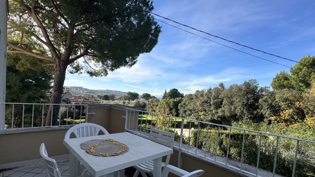 Un balcon avec une table et des chaises, une vue sur les arbres et les collines, et un ciel dégagé.