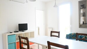 Une pièce avec une table blanche, des chaises, un téléviseur posé sur un meuble blanc, un canapé bleu et un tableau encadré accroché au mur.