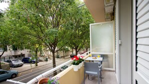 Un balcón con una mesa y sillas, plantas en macetas y vistas a una calle con coches y árboles.