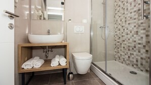 Un baño moderno con ducha envuelta en vidrio, lavamanos blanco y tocador de madera con toallas.