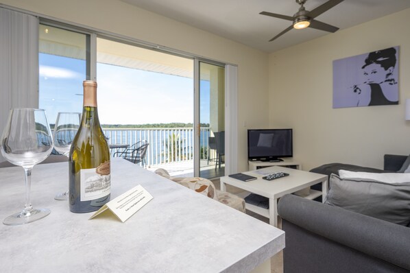 Una habitación con balcón, televisor, sofá, mesa y una botella de vino.