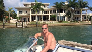 A man on a boat near a large, multi-story house with a waterfront view.