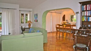 Un salon avec un canapé vert, une table à manger avec des chaises, une bibliothèque et un coin cuisine visible à travers une arche.