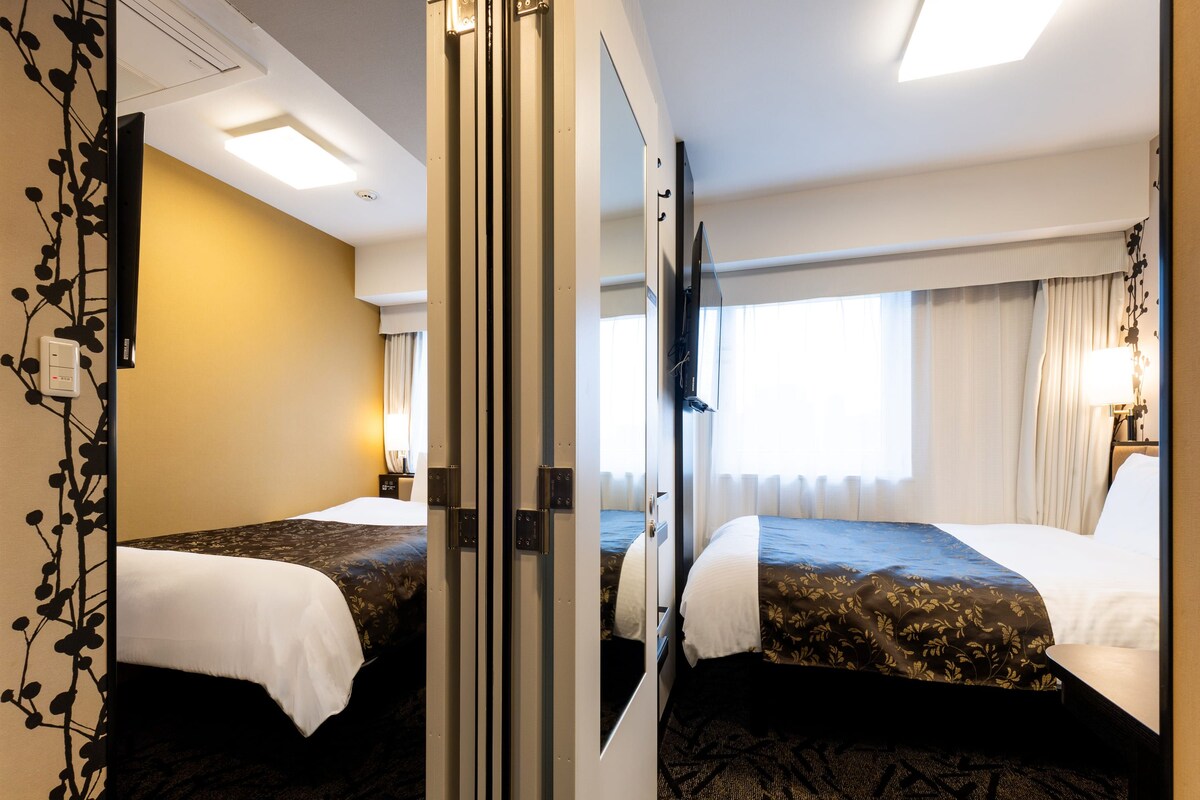 Une chambre d’hôtel avec deux lits, un grand miroir, un téléviseur à écran plat et une fenêtre avec des rideaux.