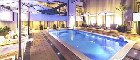 An indoor swimming pool with lounge chairs and umbrellas, surrounded by wooden decking and potted plants.