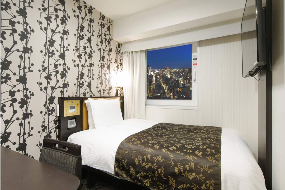 Ein Hotelzimmer mit Bett, Schreibtisch, Stuhl, Fernseher und einem Fenster mit Blick auf die Stadt.