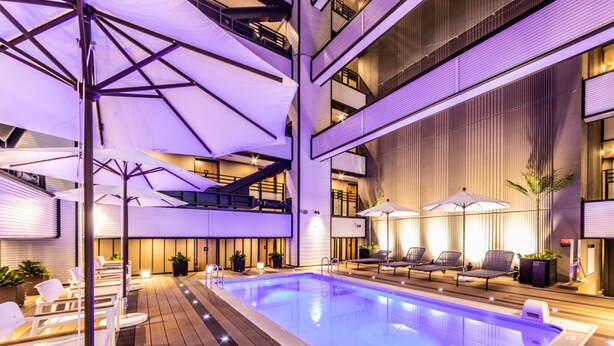 Una piscina interior con sillas de descanso y sombrillas, rodeada por una terraza de madera y arquitectura moderna.