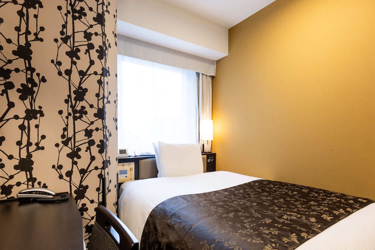 Ein Hotelzimmer mit Bett, Schreibtisch mit Telefon, Nachttisch mit Lampe und einem Fenster mit Vorhängen.