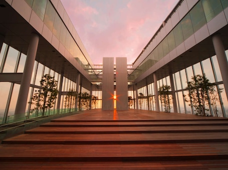 A modern building with a central vertical structure, large glass windows, and a wooden staircase leading to the entrance.