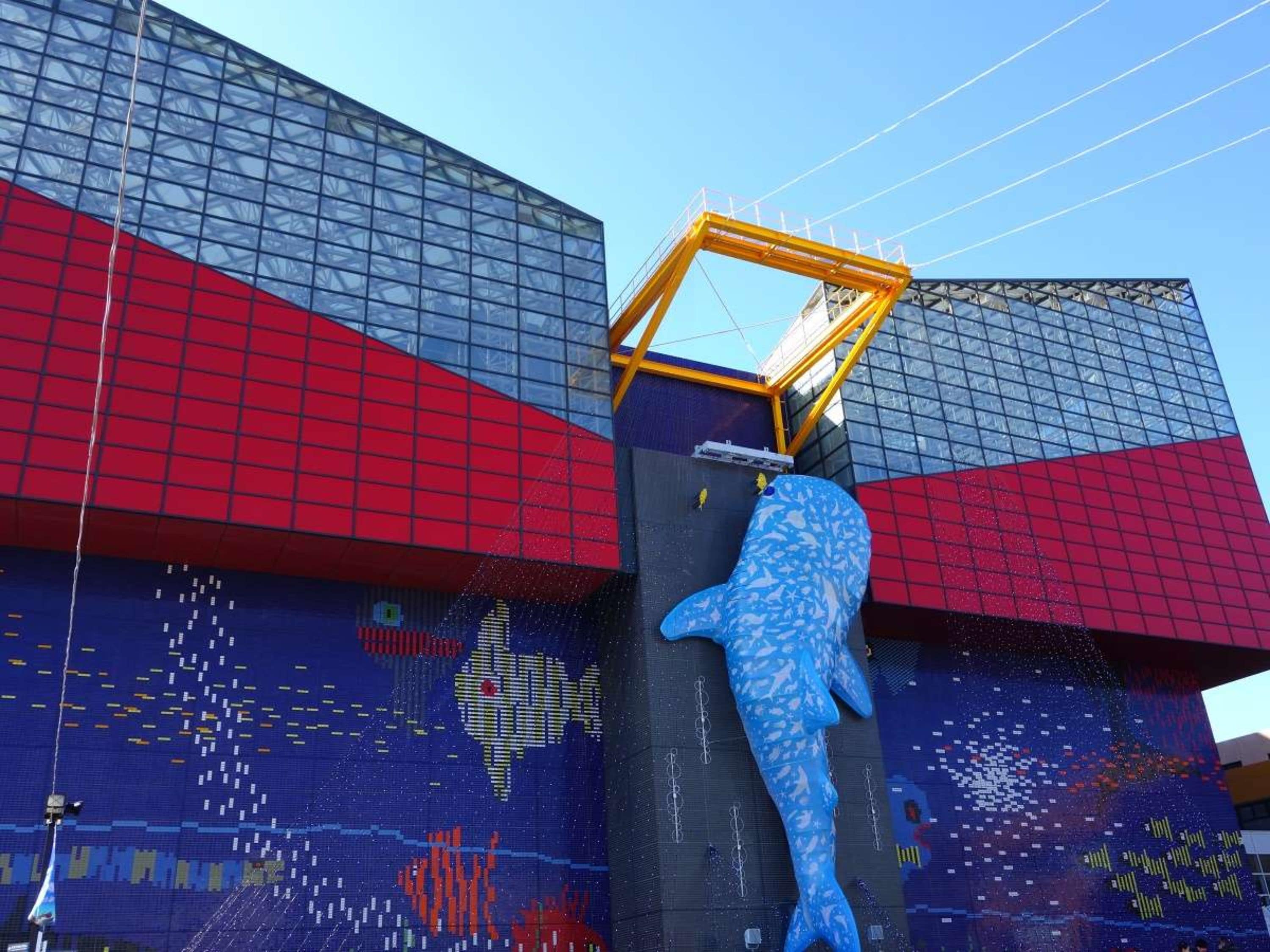 A large blue whale sculpture attached to a building with a grid-like facade and a yellow structure on top.
