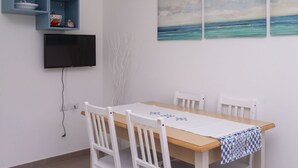 Et spiseområde med et træbord, hvide stole, en blå reol med køkkenudstyr, et fladskærms-tv og en væg med tre billeder.