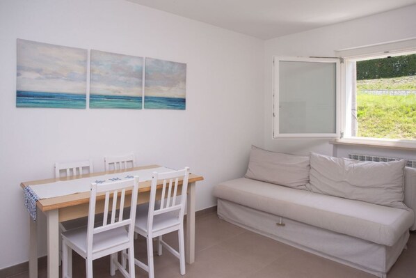 Une pièce avec une table à manger en bois, des chaises blanches, un canapé blanc et une fenêtre donnant sur un paysage verdoyant.