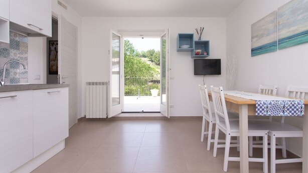 Et moderne køkken med hvide skabe, et spiseområde med hvide stole, og udsigt gennem en åben dør til en balkon.