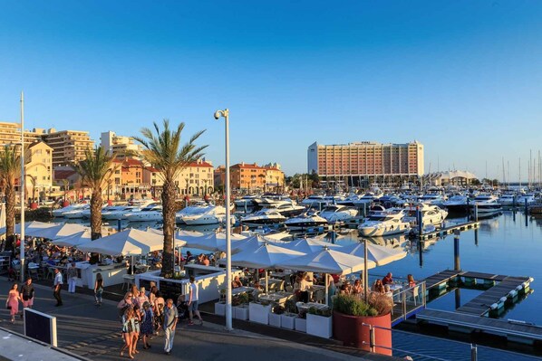 A marina with numerous boats docked, a hotel in the background, and outdoor seating areas with umbrellas.