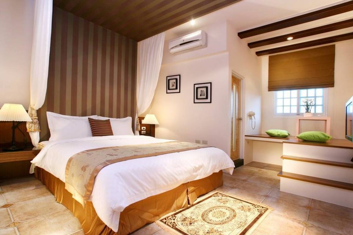 A bedroom with a large bed, bedside tables, a nightstand, an air conditioning unit, a window with a blind, and a small seating area with a bench and cushions.