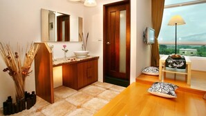 Habitación de hotel con piso de madera, un mueble de madera con lavamanos, un espejo, un televisor, una mesa con lámpara y vistas a la vegetación por la ventana.