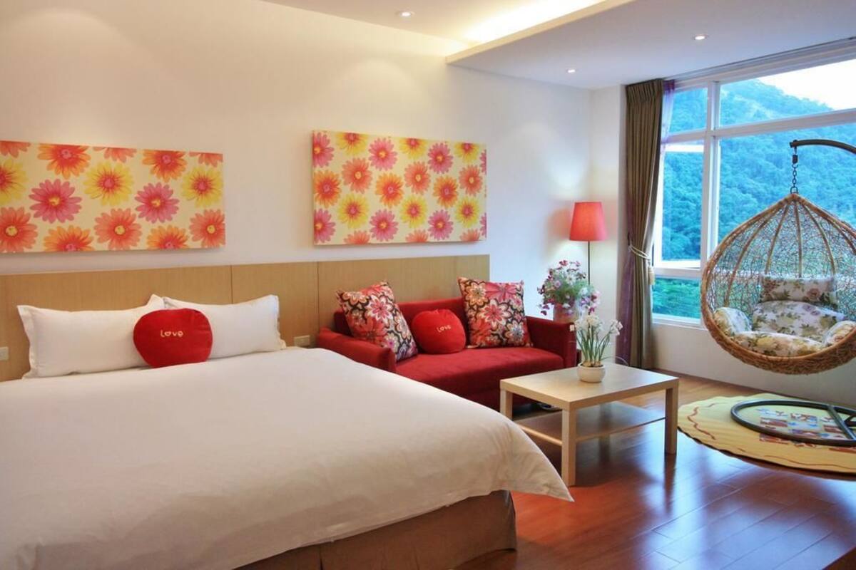 A hotel room with a bed, a red sofa, a hanging chair, and a window with a view of greenery.
