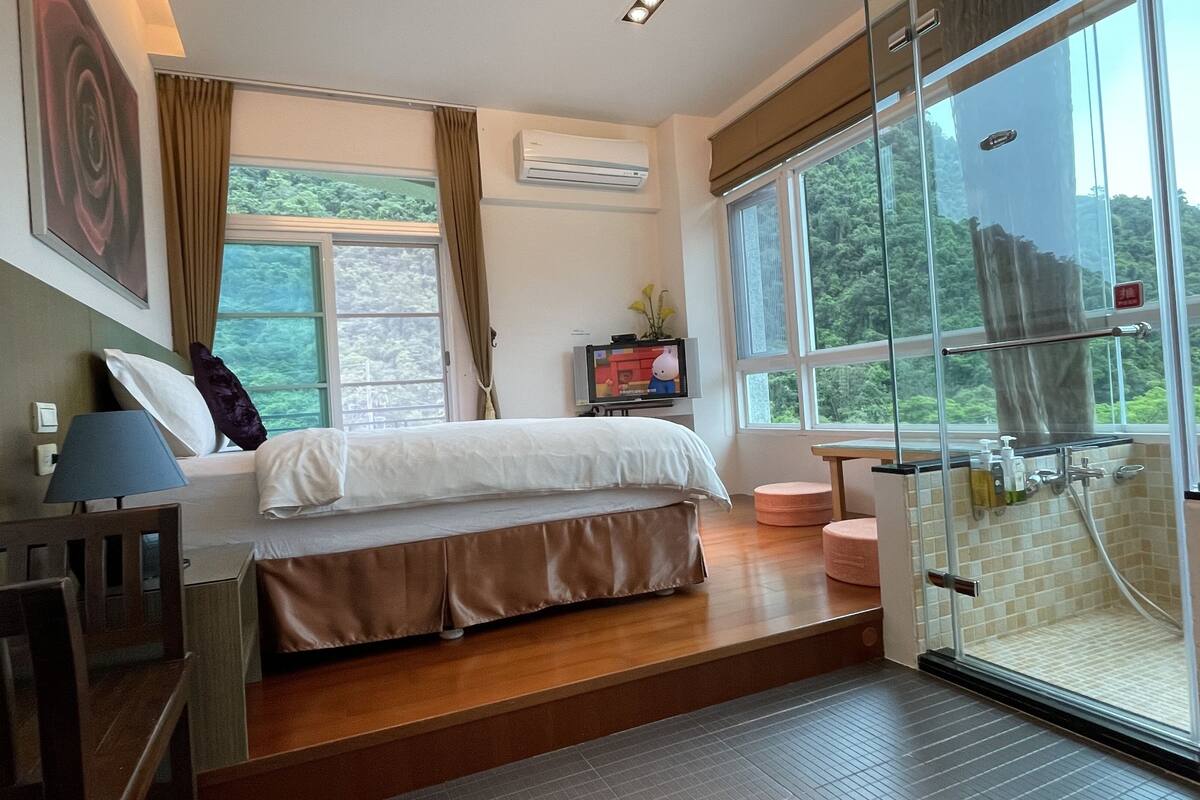 A bedroom with a bed, a TV, a window, and a shower area.