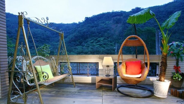 Una terraza de madera con un columpio, una silla de mimbre, una mesita con una lámpara y una planta en maceta.