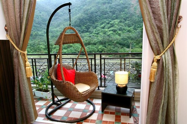 Una silla colgante de mimbre con un cojín rojo en un balcón con piso a cuadros y vista a un área boscosa.
