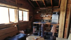 A rustic cabin interior with a wooden bench, a round wooden table, a kitchen area with a microwave, and a window with curtains. - Rustic Barn Cabin  (Pucon)