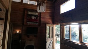 A wooden cabin interior with a fireplace, a ceiling light, and a window overlooking a deck. - Rustic Barn Cabin  (Pucon)