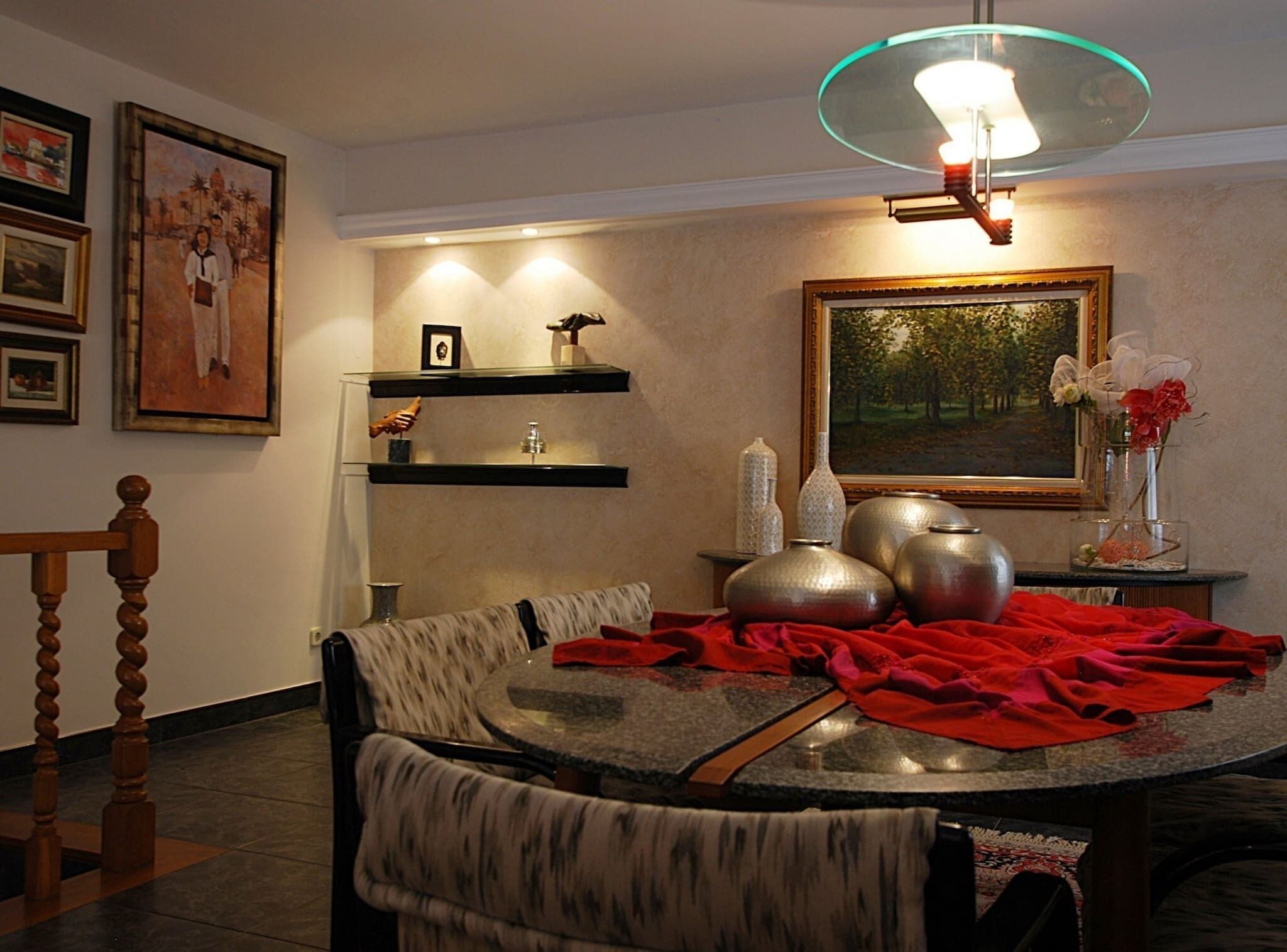 Un comedor con una mesa redonda, sillas y una araña. Hay estanterías con objetos decorativos y un cuadro en la pared.