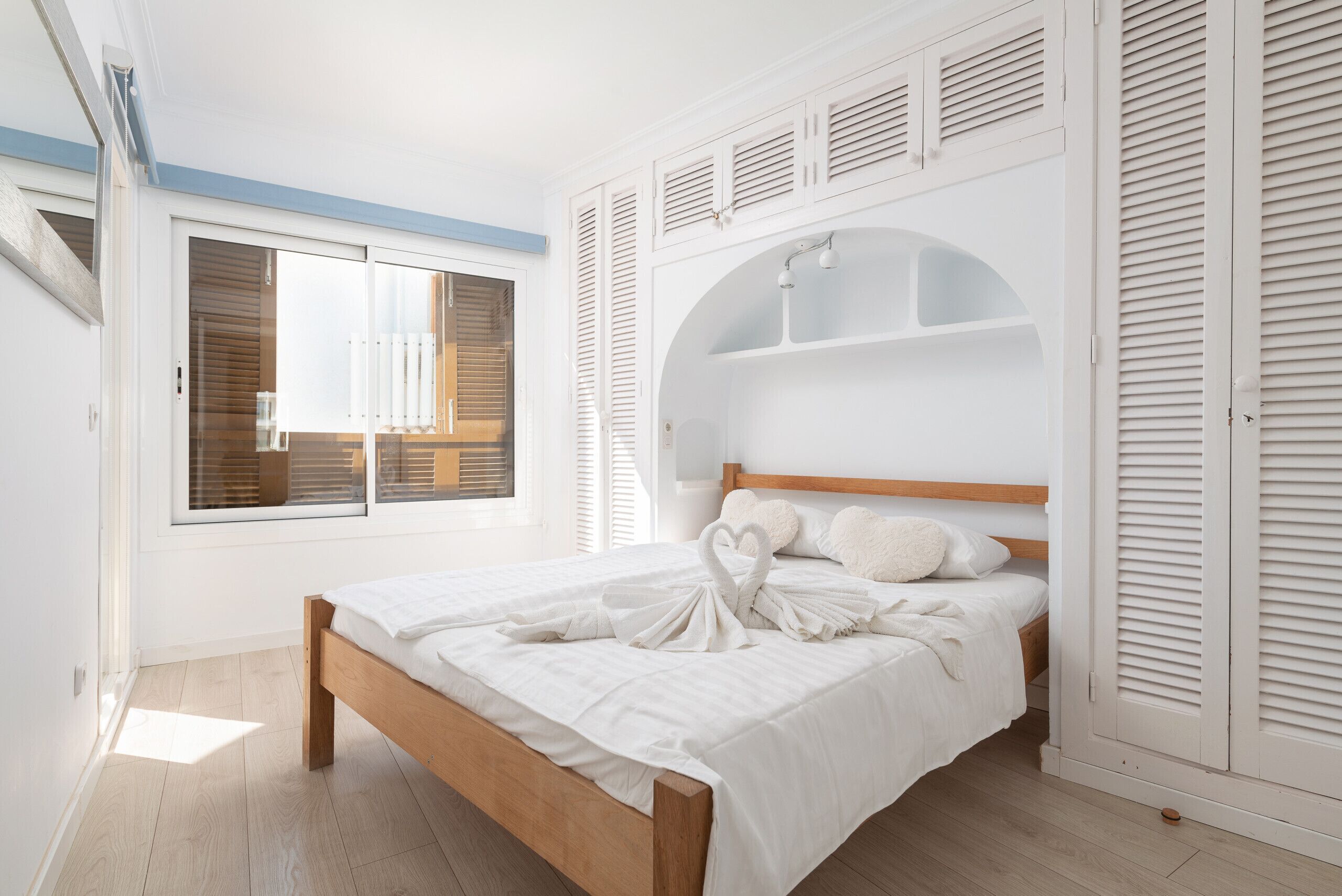 A bedroom with a wooden bed, white bedding, a window with blinds, and a built-in shelving unit above the bed.