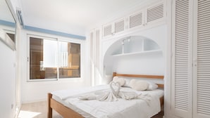 A bedroom with a wooden bed, white bedding, a window with blinds, and a built-in shelving unit above the bed. - Apartment Thierry 10 m from the beach, 2 Swimmingpools, for 4 People (Alcúdia)