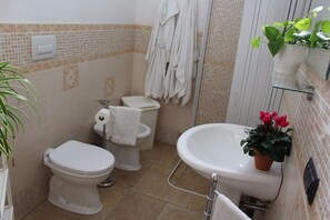 Une salle de bain équipée de toilettes, d’un bidet et d’un lavabo. Des serviettes sont accrochées au mur et une plante en pot est posée sur le lavabo.