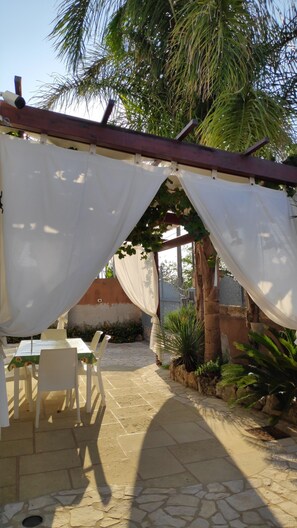 Un espace patio extérieur avec une table et des chaises sous une tonnelle blanche, entouré d’un sol en pierre et de verdure.