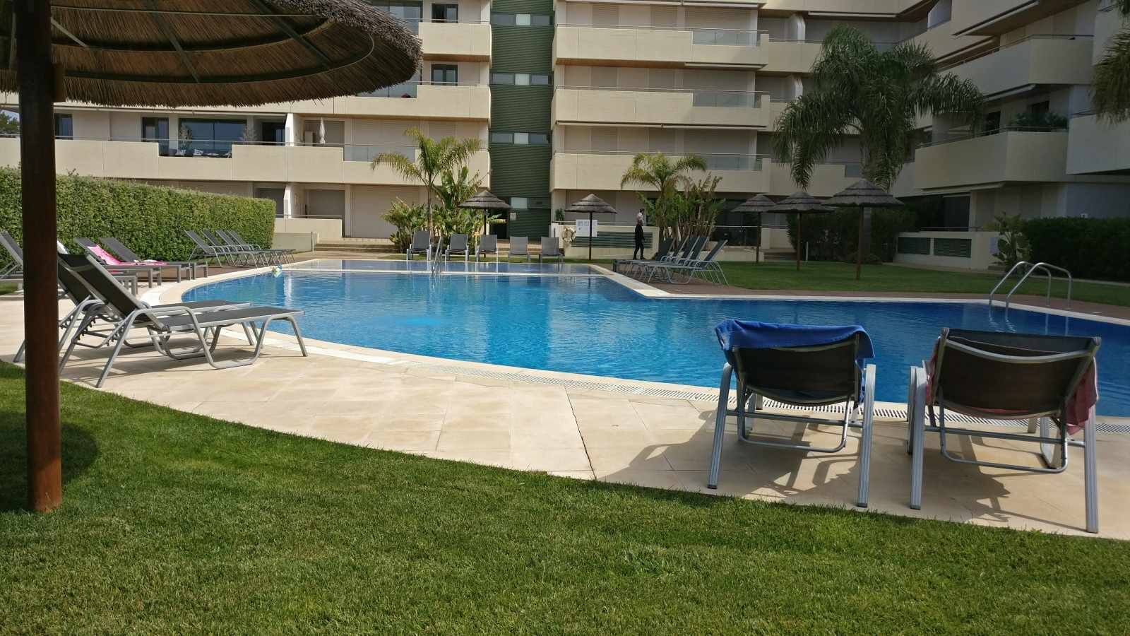 Ein Schwimmbad mit Liegestühlen und Sonnenschirmen, umgeben von einer gepflegten Rasenfläche und einem mehrstöckigen Gebäude im Hintergrund.