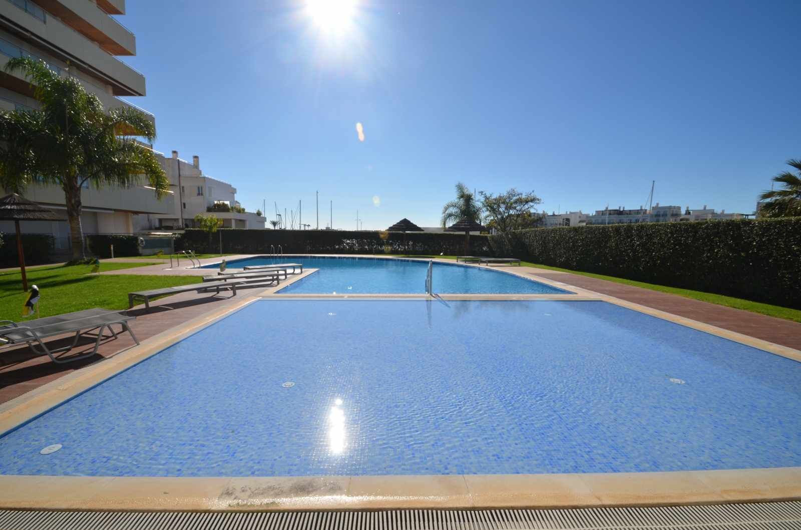 A large outdoor swimming pool with clear blue water, surrounded by a paved deck and lounge chairs.