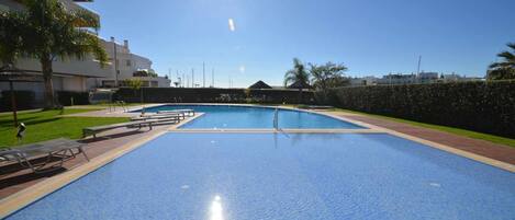 A large outdoor swimming pool with clear blue water, surrounded by a paved deck and lounge chairs.