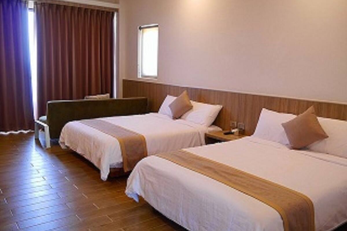 A hotel room with two beds, wooden flooring, a window with curtains, and a bedside table with a lamp.