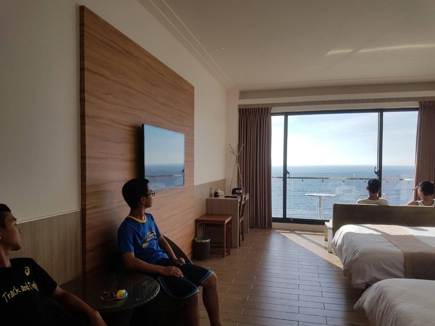A hotel room with a large window offering a view of the sea, a bed, a desk, and a television mounted on the wall.