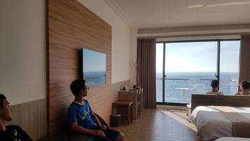 A hotel room with a large window offering a view of the sea, a bed, a desk, and a television mounted on the wall.