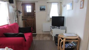 A cozy living room with a red sofa, a television, a small wooden table, and a bookshelf. - The Stable at Rambler Cottage (Eastleigh)
