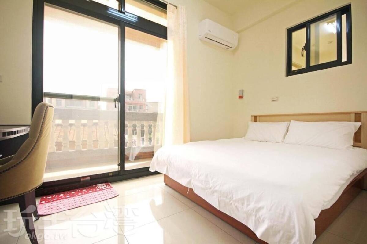 A hotel room with a bed, a chair, a window with a view of buildings, and an air conditioning unit.