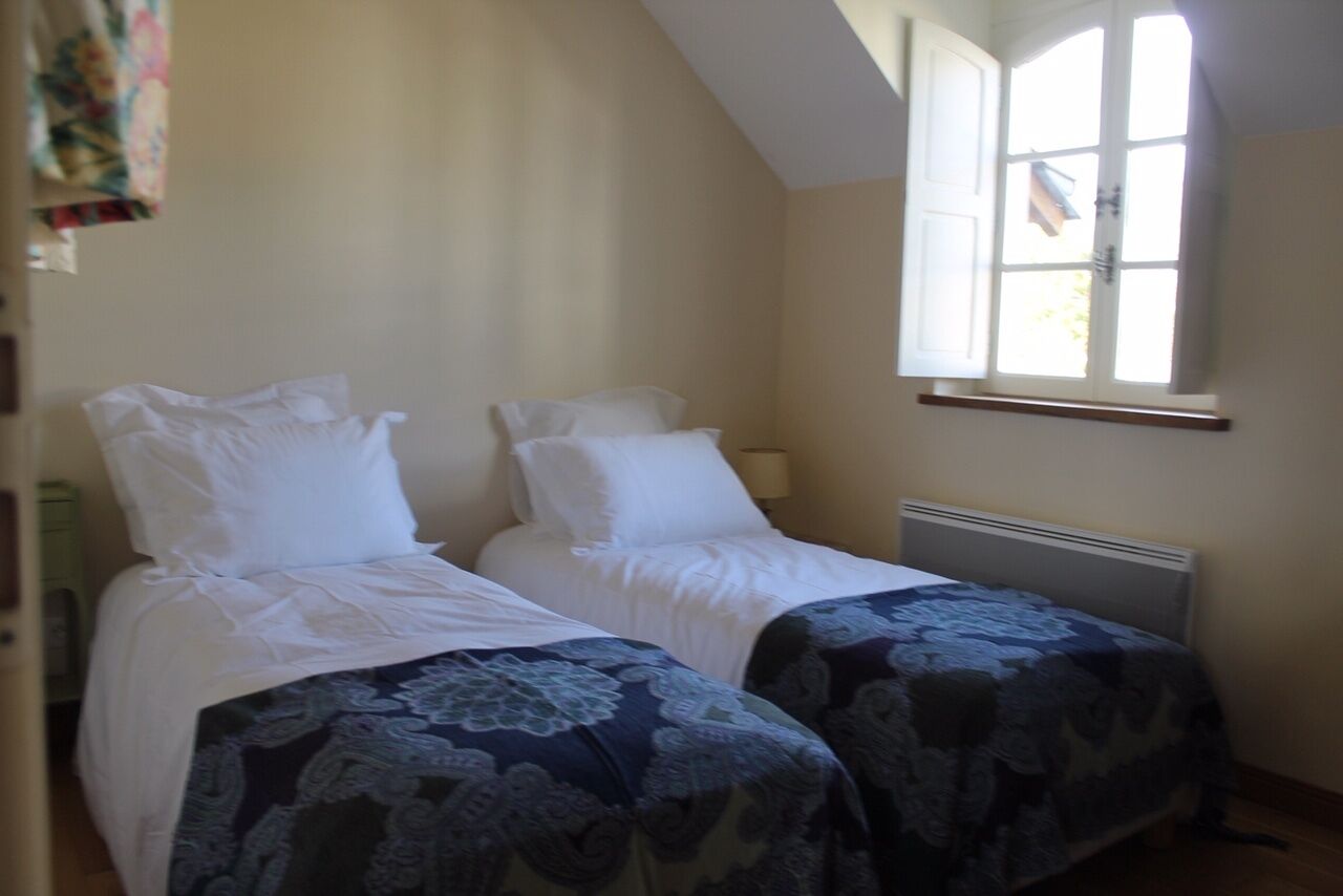 Une chambre avec deux lits, chacun recouvert d’une literie blanche et d’une couvre-lit à motifs floraux d’un côté. Il y a une fenêtre avec des volets blancs et un radiateur en dessous.