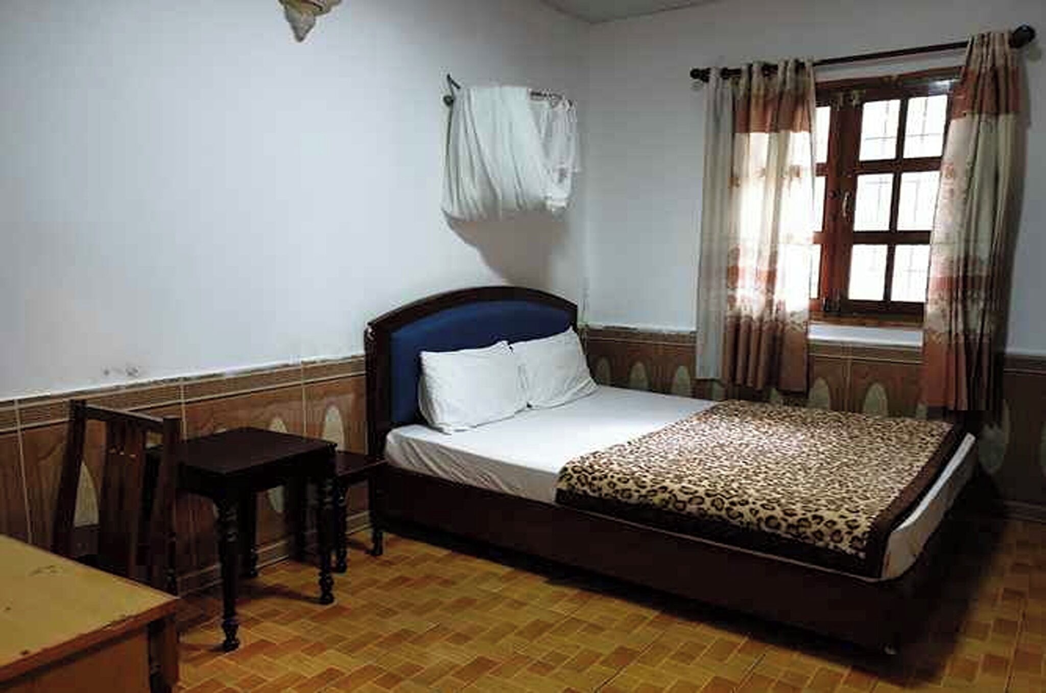 A bedroom with a bed, a small table, a window with curtains, and a wall-mounted towel rack.