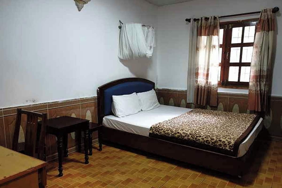 Ein Schlafzimmer mit Bett, einem kleinen Tisch, einem Fenster mit Vorhängen und einem an der Wand befestigten Handtuchhalter.