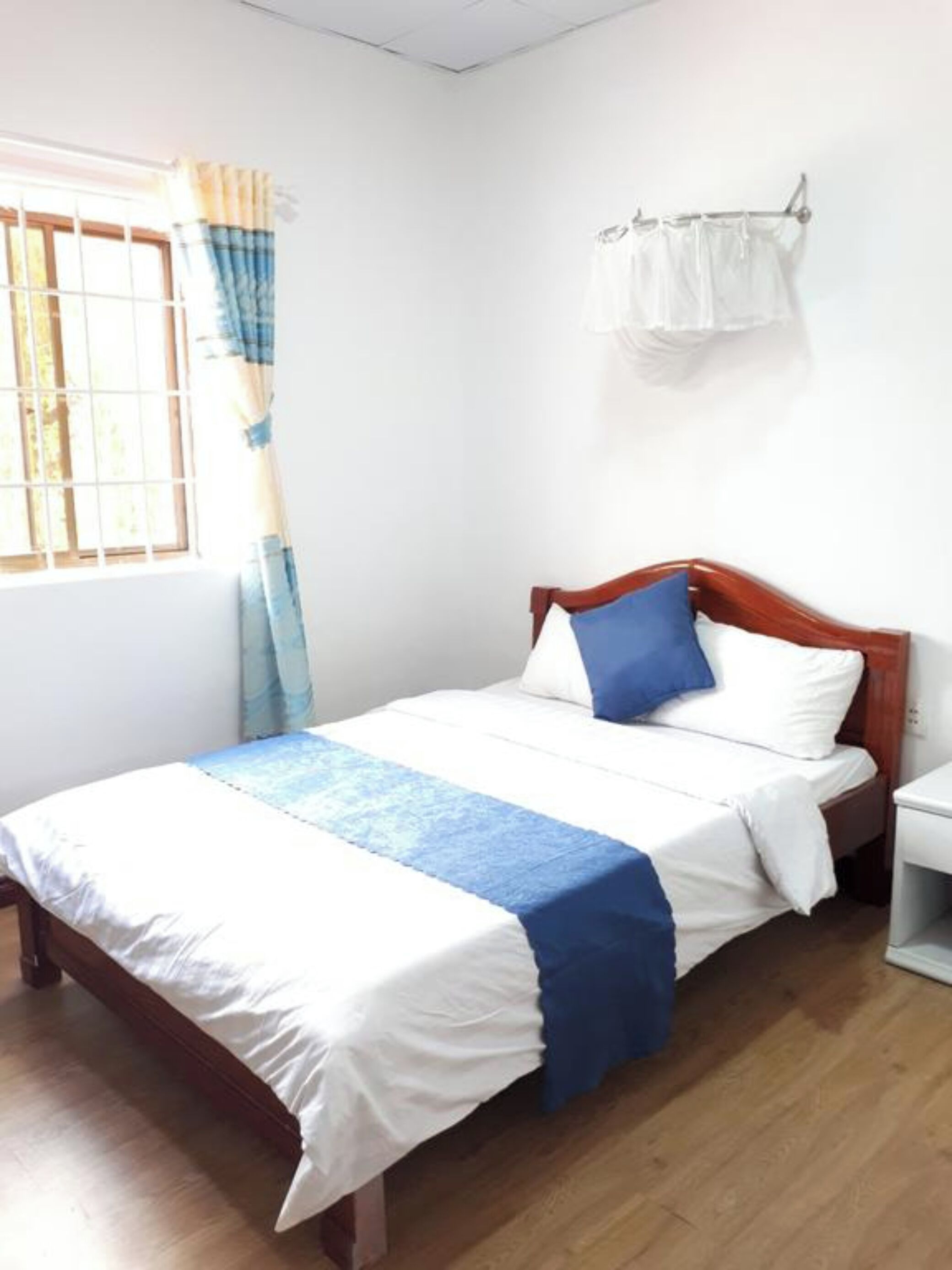 A single bed with a wooden headboard, a white nightstand, a window with curtains, and a wall-mounted light fixture.