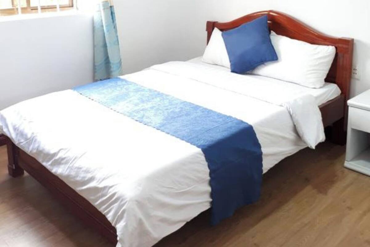 Un lit simple avec une tête de lit en bois, une table de chevet blanche, une fenêtre avec des rideaux et un luminaire fixé au mur.