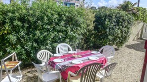 An outdoor dining area with a table set for six, surrounded by white plastic chairs, against a backdrop of dense green foliage. - Jean Michel by Interhome (Capbreton)
