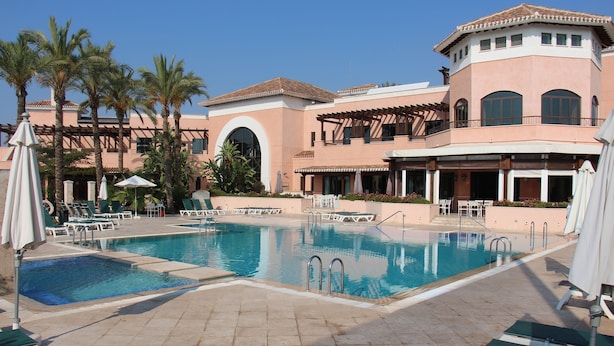 A pool area with multiple lounge chairs and umbrellas, surrounded by palm trees and a building with a Mediterranean architectural style.