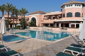 A pool area with multiple lounge chairs and umbrellas, surrounded by palm trees and a building with a Mediterranean architectural style. - Villa With Pool, Air Conditioning and WiFi on a private 18 hole golf course. (Torre-Pacheco)