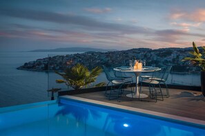 A poolside dining area with a table and chairs overlooking a coastal cityscape at dusk. - Luxury Villa Riva with Infinity Pool (Okrug Gornji)